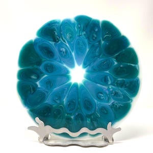 May include: A round, decorative plate with a swirling pattern of teal, turquoise, and white resin. The plate is displayed on a silver-colored metal stand with a wavy design. The plate's design resembles a cross-section of a citrus fruit.