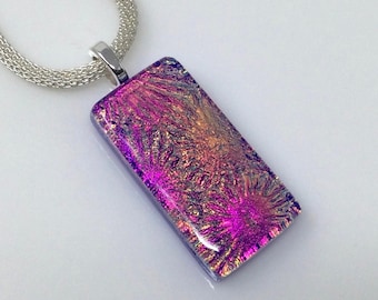 May include: A rectangular pendant necklace with a dichroic glass design. The pendant has a vibrant mix of pink, purple, and gold colours. The necklace has a silver chain and a silver bail.