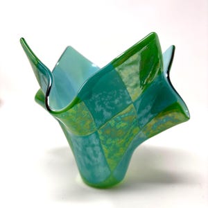 May include: A decorative glass bowl with a unique, ruffled shape. The bowl features a checkerboard pattern in shades of green and blue, with a glossy finish. The edges are gently curved, adding to its artistic appeal.