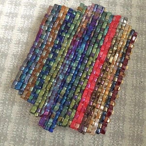 May include: A woven coaster with a rectangular shape. The coaster is made of small, iridescent, rectangular beads in various colors, including blue, green, purple, and gold. A vertical stripe of red beads runs down the center.