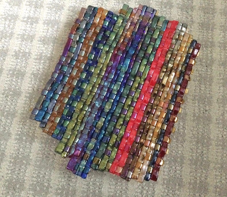 May include: A woven coaster with a rectangular shape. The coaster is made of small, iridescent, rectangular beads in various colors, including blue, green, purple, and gold. A vertical stripe of red beads runs down the center.