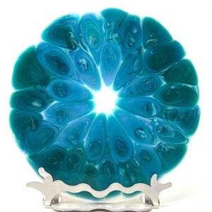 May include: A round, decorative plate with a swirling blue and teal pattern, resembling a flower or abstract design. The plate is displayed on a silver-colored metal stand, creating a unique display piece.