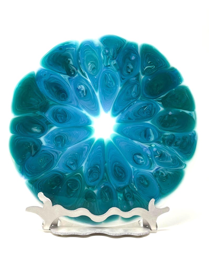 May include: A round, decorative plate with a swirling blue and teal pattern, resembling a flower or abstract design. The plate is displayed on a silver-colored metal stand, creating a unique display piece.