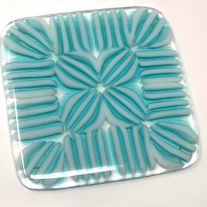 May include: Square glass coaster with a turquoise and white striped design. The coaster features a repeating pattern of curved and straight lines, creating a decorative and functional item. The glass is clear, allowing the design to be visible.