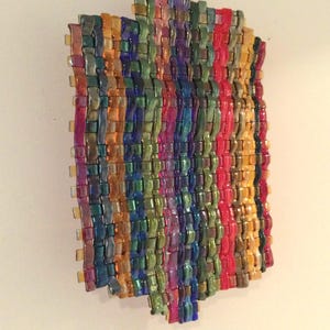May include: A colorful, woven glass art piece. The artwork features a rectangular shape with vertical stripes of various colors, including red, yellow, green, blue, and purple. The glass has a glossy finish, creating a vibrant and textured appearance.