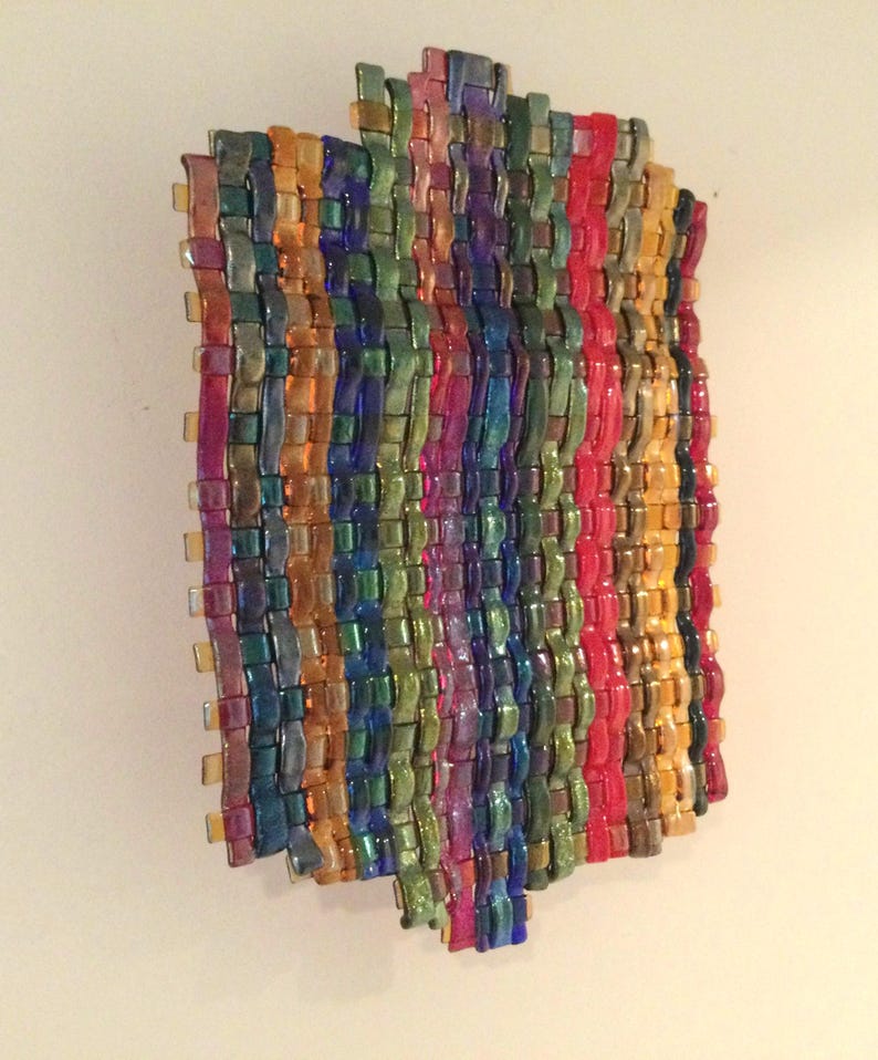 May include: A colorful, woven glass art piece. The artwork features a rectangular shape with vertical stripes of various colors, including red, yellow, green, blue, and purple. The glass has a glossy finish, creating a vibrant and textured appearance.
