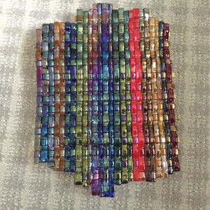 May include: A rectangular, woven piece composed of small, iridescent beads in a variety of colors. The beads are arranged in vertical rows, with a prominent red stripe down the center. The color palette includes blues, greens, purples, and golds.