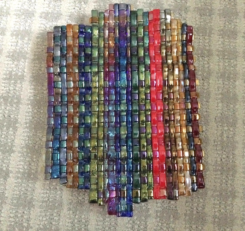 May include: A rectangular, woven piece composed of small, iridescent beads in a variety of colors. The beads are arranged in vertical rows, with a prominent red stripe down the center. The color palette includes blues, greens, purples, and golds.