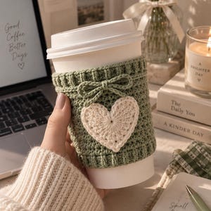 Puede incluir: Un vaso de café reutilizable blanco con una funda de ganchillo en color verde salvia. La funda está decorada con un pequeño lazo de ganchillo y un corazón central de color crema. Se aprecia la textura del hilo con bordes acanalados.