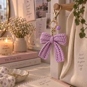 May include: A lavender crochet bow keyring featuring a gold-toned metal lobster clasp, a circular ring, and a small matching purple bead. The bow is constructed from yarn with a visible textured stitch pattern, hanging from a wooden hook.