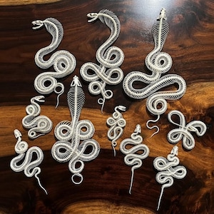 May include: A collection of ten white, articulated snake skeleton figures in various sizes and coiled positions. Each piece features a detailed skull, a long spine, and rib-like structures that create a flexible, segmented appearance.
