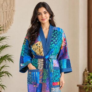 Op de afbeelding: Een knielange kamerjas met een levendig patchworkontwerp met diverse blauwe, turquoise en veelkleurige bloemen- en geometrische patronen. Het kledingstuk heeft lange mouwen, een effen blauwe kraag, een bijpassende ceintuur en twee voorzakken.