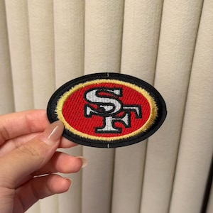 Puede incluir: Un parche bordado de forma ovalada con el logotipo de los San Francisco 49ers. El diseño incluye un fondo rojo con un borde dorado, un borde exterior negro y las letras SF en blanco con contornos negros y un efecto de sombra paralela.