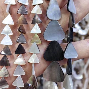 May include: Multiple strands of polished mother-of-pearl beads shaped like triangles. The beads feature a variety of iridescent colours including white, grey, charcoal, and brown, with smooth surfaces and a natural lustre arranged in vertical rows.