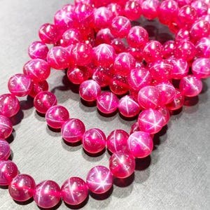 May include: A strand of round, translucent fuchsia-pink beads, each featuring a distinct six-rayed star effect known as asterism. The polished, spherical beads are arranged in a loose, coiled formation, highlighting the bright pink color.