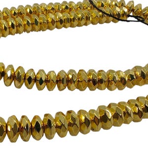May include: A long strand of metallic gold-coloured beads with a faceted, rondelle-shaped design. The beads are strung together on a thin black thread, creating a continuous loop of reflective, shiny surfaces that catch the light uniformly.