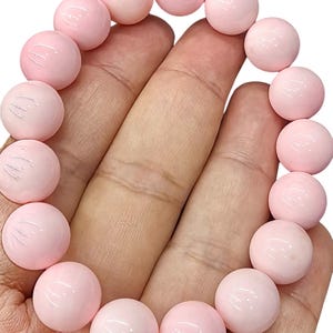 以下が含まれることがあります： 滑らかで丸いライトピンクのビーズで構成されたストレッチブレスレット。ビーズはサイズと形が均一で、光を反射する光沢のある仕上げが特徴です。ブレスレットは円形に配置されており、ジュエリー全体を通して一貫したパステルピンクの色合いを示しています。