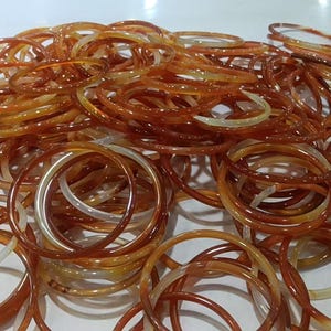 May include: A large collection of thin, circular bangles in varying shades of translucent amber, orange, and cream. The bracelets feature a smooth, polished finish with swirling color patterns throughout each individual ring-shaped piece.