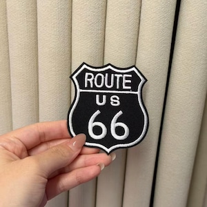 Könnte beinhalten: Ein schwarz-weiß bestickter Aufnäher in Schildform mit dem Text ROUTE US 66 in weißen Buchstaben. Der Aufnäher hat einen markanten schwarzen Hintergrund mit einem weißen Rand, der das Design des ikonischen Straßenschildes nachahmt.
