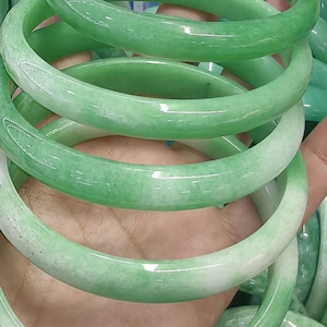 May include: A collection of smooth, polished jade-style bangles featuring a rounded profile. Each bangle displays a translucent appearance with varying gradients of vibrant green and milky white marbling throughout the circular stone bands.
