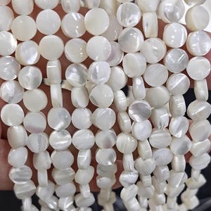 Puede incluir: Múltiples hilos de cuentas de nácar blanco, cada una con forma de pequeño disco circular plano. Las cuentas presentan un brillo natural iridiscente y están ensartadas en filas verticales, mostrando una textura de superficie lisa y pulida.