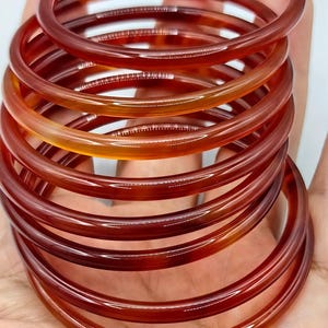 May include: A stack of multiple smooth, circular bangles made of translucent, polished stone in varying shades of deep orange, reddish-brown, and amber. The bangles have a rounded profile and a glossy finish, showing natural color variations.
