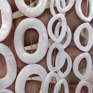 May include: A collection of white mother-of-pearl beads shaped like oval rings, strung together on thin clear filaments. The beads feature a smooth, iridescent surface with a glossy finish, arranged in multiple vertical strands of varying sizes.