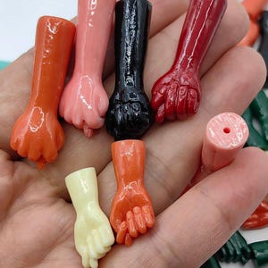 Puede incluir: Una colección de pequeñas cuentas brillantes con forma de puño en varios colores, incluyendo naranja, rosa, negro, rojo y crema. Cada cuenta presenta una parte superior cilíndrica con un orificio para enhebrar y una base con forma de mano.
