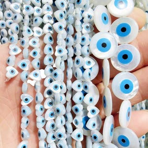 May include: A collection of white mother-of-pearl beads in heart and circular shapes, each featuring a central blue and black eye design. These smooth, iridescent beads are arranged in multiple vertical strands, showcasing the eye motif.