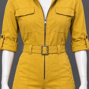 May include: A mustard yellow utility jumpsuit featuring a collared neckline, a full-length front zipper, and two chest pockets. The garment includes three-quarter length sleeves with buttoned tabs, a matching fabric belt with a buckle, and cargo pockets.