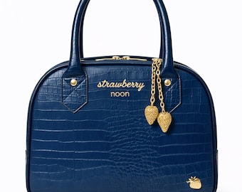 Puede incluir: Un bolso de mano azul oscuro con textura de cocodrilo, con dos asas superiores y herrajes dorados. El frente muestra el texto "strawberry noon" en letras doradas, junto con dos dijes de fresa dorados y un pequeño emblema de fresa.