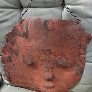 Può includere: Un oggetto da appendere alla parete color terracotta che raffigura un volto umano scolpito con occhi chiusi, naso e labbra. Il pezzo presenta un bordo organico irregolare ed è sospeso tramite una sottile catena in metallo scuro fissata in alto.