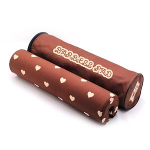 May include: Two cylindrical barbell pads in a dark brown color. One pad features a repeating pattern of small cream-colored hearts. The other pad displays the text BARBELL PAD in a cream-colored font with a decorative outline on its side.