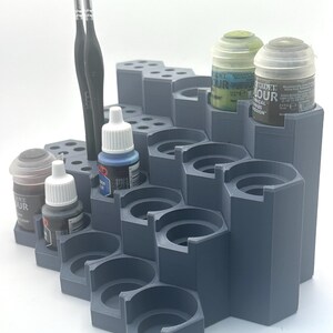 May include: A tiered gray plastic paint organizer with a hexagonal grid structure. The stand features multiple circular cutouts for holding paint pots and small brush slots. Several Citadel brand paint pots and two thin brushes are inserted.