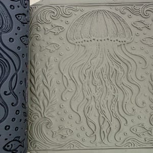 May include: A dark gray cylindrical rolling pin with a recessed jellyfish, fish, and seaweed pattern next to a light gray square clay tile featuring the same embossed marine design of a central jellyfish surrounded by fish, bubbles, and plants.