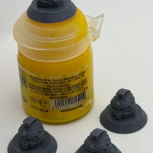 May include: A yellow paint bottle with a 0.6 fl oz capacity and three dark gray 3D-printed miniature helmets on circular bases. One helmet is placed on the bottle cap, while two others sit separately. The label includes text about Games Workshop.