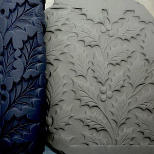 Könnte beinhalten: Eine dunkelblaue, zylindrische Ton-Texturwalze mit einem erhabenen Muster aus Stechpalmenblättern und Beeren, platziert neben einer flachen, grauen Tonplatte, die mit demselben detaillierten botanischen Design auf ihrer Oberfläche geprägt ist.