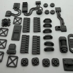 Könnte beinhalten: Eine Sammlung grauer, 3D-gedruckter Kunststoff-Miniaturbauteile für den Modellbau. Das Set enthält verschiedene mechanische Teile wie rechteckige Paneele mit Gittermustern, gebogene Rohre, kreisförmige Lüftungsschlitze, kleine Griffe und Strukturrahmen.