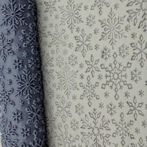 May include: A cylindrical grey rolling pin with a raised snowflake pattern positioned next to a flat, light-coloured surface that displays the same snowflake design pressed into it, creating a repeating winter-themed texture across the material.