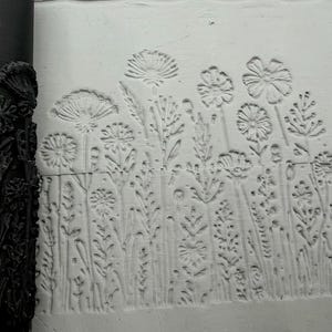 Könnte beinhalten: Eine schwarze zylindrische Teigrolle mit erhabenem Blumenmuster liegt neben einer weißen Tonoberfläche, in die dasselbe Blumendesign eingedrückt ist. Das Muster zeigt verschiedene Wildblumen, Stängel und Blätter im Relief.