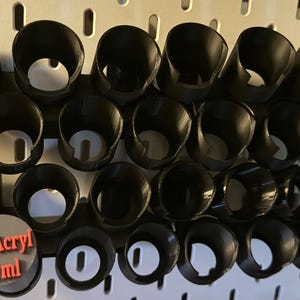 以下が含まれることがあります： 塗料ボトル用の複数の円形スロットを備えた黒いプラスチック製収納ラック。前面の隅には「ProAcryl 22 ml」という赤い文字が書かれた灰色の円形ラベルが付いており、白い有孔ボード壁パネルに取り付けられるように設計されています。