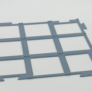 Op de afbeelding: Een platte, grijze, 3D-geprinte rasterstructuur met een drie-bij-drie opstelling van negen vierkante openingen. Het frame heeft in elkaar grijpende lipjes langs de buitenranden om het met andere delen te verbinden.