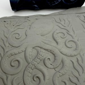 Op de afbeelding: Een donkerblauwe cilindrische patroonroller geplaatst boven een platte plaat grijze klei. De klei vertoont een diep ingedrukt ontwerp van een octopus met kronkelende tentakels en zeewier, passend bij het patroon op de roller.