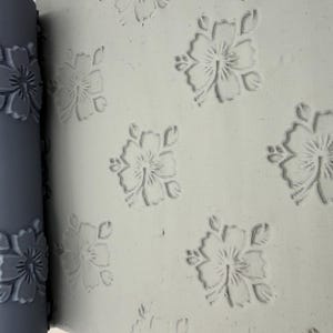Könnte beinhalten: Ein zylindrisches graues Nudelholz mit erhabenem Blumenmuster liegt neben einer flachen, hellgrauen Tonplatte. Das Nudelholz weist ein sich wiederholendes, hibiskusartiges Blumenmuster auf, das in die Oberfläche der Tonplatte eingedrückt ist.