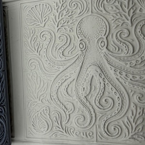 Puede incluir: Un rodillo cilíndrico de color gris oscuro con un patrón de pulpo y conchas en relieve junto a una losa de arcilla gris claro que presenta el mismo diseño grabado de un pulpo central rodeado de algas y varios motivos de conchas.