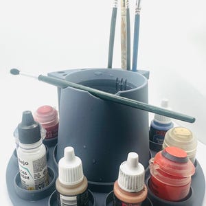 May include: A dark grey plastic paint brush and paint bottle organiser featuring a central water basin with integrated brush slots. The circular base holds multiple small paint bottles of various colours, while several paint brushes are stored upright.