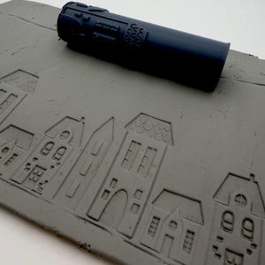 May include: A dark blue cylindrical rolling pin with a textured pattern sits atop a flat grey slab of clay. The clay slab features an embossed design of a row of houses with various roof shapes, windows, and doors imprinted into the surface.