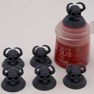 May include: Five dark gray 3D-printed horned skulls on circular bases, with one additional horned skull resting on top of a red Citadel paint pot labeled 0.4 fl oz. The skulls feature visible horizontal print layers and hollow eye sockets.