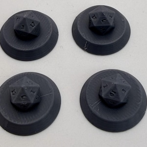 May include: Four black 3D-printed gaming accessories, each consisting of a circular base with a twenty-sided die shape integrated into the center. The objects feature a visible layered texture from the printing process and numerical markings.