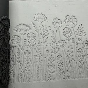 Könnte beinhalten: Eine schwarze zylindrische Tonrolle mit einem komplizierten geprägten Blumenmuster aus verschiedenen Wildblumen und Stängeln, neben einer hellgrauen Tonplatte, die denselben detaillierten botanischen Abdruck im Relief zeigt.